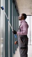 Black man professional cleaning service worker in overalls cleans the pvc windows in office. Vertical video