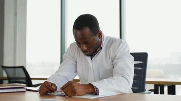 Stressed frustrated black doctor in white uniform with headache sitting alone at work desk in hospital. Sad young gp feeling desperate and guilty for medical failure video