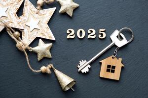 House key with keychain cottage on festive black background with stars, lights of garlands. New Year 2025 wooden letters, greeting card. Purchase, construction, relocation, mortgage, insurance photo