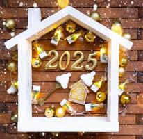 House key with keychain cottage on festive brown wooden background with stars, lights of garlands. New Year 2025 golden letters under the roof. Purchase, construction, relocation, mortgage, insurance photo