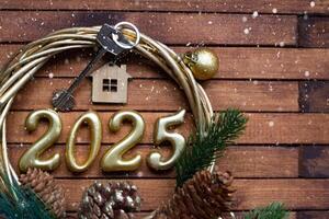 New Year House key with keychain cottage on festive brown wooden background with number 2025 in wreath, lights of garlands. Purchase, construction, relocation, mortgage, insurance photo