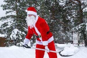 Santa Claus cleans snow with shovel in winter outdoors after a snowfall. Cleaning the streets in the village, clearing the passage for cars, difficult weather conditions for Christmas and New Year photo