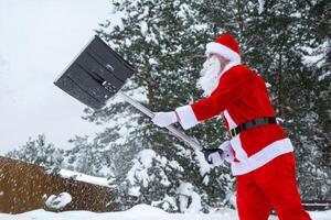 Santa Claus cleans snow with shovel in winter outdoors after a snowfall. Cleaning the streets in the village, clearing the passage for cars, difficult weather conditions for Christmas and New Year photo