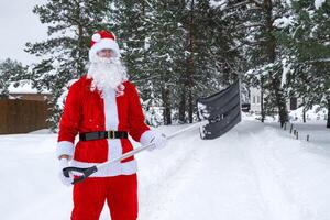 Santa Claus cleans snow with shovel in winter outdoors after a snowfall. Cleaning the streets in the village, clearing the passage for cars, difficult weather conditions for Christmas and New Year photo