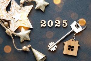 House key with keychain cottage on festive black background with stars, lights of garlands. New Year 2025 wooden letters, greeting card. Purchase, construction, relocation, mortgage, insurance photo