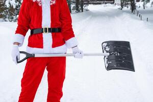 Santa Claus cleans snow with shovel in winter outdoors after a snowfall. Cleaning the streets in the village, clearing the passage for cars, difficult weather conditions for Christmas and New Year photo
