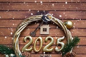 New Year House key with keychain cottage on festive brown wooden background with number 2025 in wreath, lights of garlands. Purchase, construction, relocation, mortgage, insurance photo