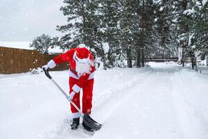Santa Claus cleans snow with shovel in winter outdoors after a snowfall. Cleaning the streets in the village, clearing the passage for cars, difficult weather conditions for Christmas and New Year photo