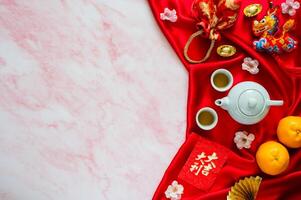 Tea set on red cloth background with red envelope packets or ang bao word mean auspice, red bag, ingots word mean wealth, hanging pendant dragon and oranges on marble table. photo