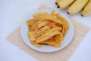 crujiente plátano papas fritas en un blanco plato con Fresco plátanos foto