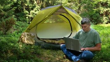 A man works on a laptop in a tourist camp in a beautiful forest. Concept of freelancing, digital nomad or remote office. 4K video