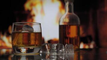 Glass and bottle of whiskey, scotch or bourbon stands on a table against background of a fireplace with a flame. Alcoholic drink and home comfort concept. video