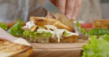 Female chef cuts club sandwich in a half by sharp knife on a wooden board at the kitchen. Big appetizing Sandwich with ham, tomato slices and pieces of cheese, lettuce and mayonnaise sauce. Ultra 4K video