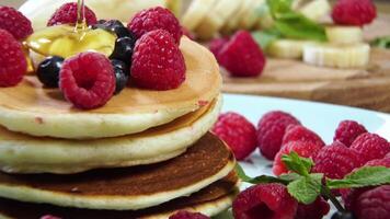 Closeup of Pouring honey on stack of pancakes. Tasty breakfast food. Pancakes are served with raspberries, banana and mint leaf. Ultra 4K video
