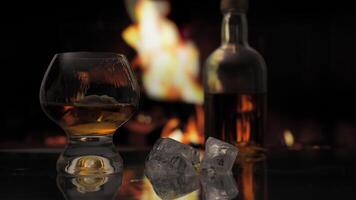 Glass of cognac, whiskey, scotch or bourbon with ice stands on a table against background of a fireplace with a flame. Alcoholic drink and home comfort concept. video