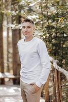 de cerca vertical retrato de un adolescente en un blanco suéter y marrón pantalones. contento sonriente adolescente en verano parque en luz de sol. un hermosa niño es mirando a el cámara en el claro. foto