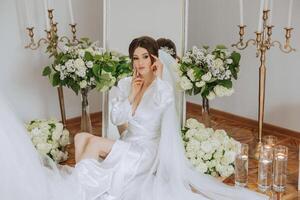 un hermosa morena novia es consiguiendo Listo para el Boda en un hermosa tocador estilo atuendo sentado en un silla entre Fresco flores y elegante candelabros Boda retrato, foto. foto