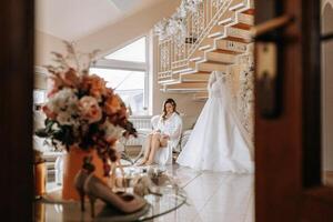 Morning of the bride before the wedding. A beautiful young woman with a veil and a beautiful hairstyle in a white robe near her wedding dress on a mannequin. Professional makeup. Last preparations. photo