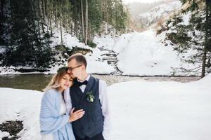 The newlyweds are out of focus against the background of a pine forest and a river. The bride in a white wedding dress is wrapped in a blue blanket. Winter wedding. photo