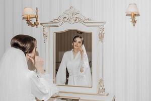 retrato de un joven hermosa novia en un hermosa hotel interior preparando para su boda. un maravilloso mujer mira en el espejo a su maquillaje y peinado foto