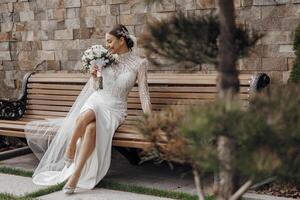 Portrait. The bride in an elegant long dress and veil, holding a bouquet, sits on a bench in the yard. Wedding in nature photo