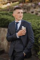 The groom in a black suit adjusts the boutonniere, poses against the background of a green tree. Wedding portrait. photo