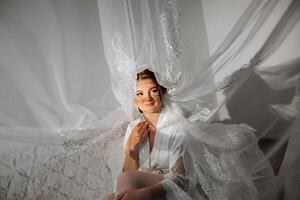 Nice cute girl in a boudoir robe. Happy beautiful bride under veil. Fashion, beauty, style. Morning of the bride. Beautiful bride with stylish make up and hair style. photo