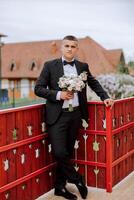 The groom in a black suit holds a bouquet, poses leaning on a red railing. Wedding portrait. photo