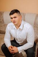 joven sonriente hombre en blanco camisa sentado en sofá mirando a cámara. un novio con un corto peinado es preparando para su Boda foto