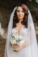 Curly brunette bride in a lace dress with an open bust, in a lush veil holds a bouquet and poses against the background of green trees. Spring wedding photo