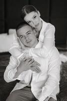 joyful young bride in white pajamas hugs the shoulders of handsome groom in white unbuttoned shirt while sitting together in modern hotel room before wedding ceremony. Black and white photo