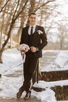 portrait of a handsome and manly groom in a black suit with a tie. Groom in nature in the winter season. photo