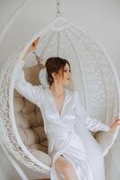 Morning of the bride in boudoir style. A woman bride in a white robe sits on a wicker swing near her wedding dress on a mannequin. Wedding day. photo