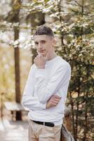 Close-up vertical portrait of a teenager in a white sweater and brown pants. Happy smiling teenager in summer park in sunlight. A beautiful child is looking at the camera in the clearing. photo