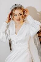 Wedding portrait. A blonde bride in a silk suit and tiara poses while sitting in a chair in her room photo