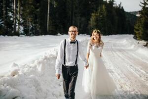 The bride and groom are running along a snowy road against the background of a pine forest and beautiful contrast sunlight. Side view. Winter wedding. Place for logo. photo