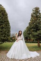 Curly brunette bride in a lush veil and dress with long sleeves, holding a bouquet and posing against a background of green trees. Spring wedding photo