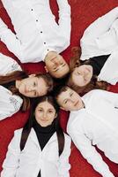 Happy friends lying together in a circle looking at the camera, holding hands and smiling while lying on a red wall... Friendship, lifestyle, togetherness, business and teamwork concepts. photo