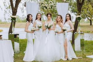 Group portrait of the bride and bridesmaids. Bride in a wedding dress and bridesmaids in white dresses and holding stylish bouquets on the wedding day against the background of the lake. photo