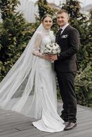 Portrait. A bride in an elegant long dress and veil and a groom in a suit pose while standing near trees. Wedding in nature photo