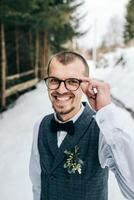 Boda retrato de el novio. el novio soportes en contra el antecedentes de el invierno bosque. un hombre en un chaleco y blanco camisa, lentes y un arco atar. invierno Boda foto