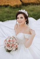 brunette bride in off-the-shoulder lace white dress and tiara posing with a bouquet of white and pink flowers while sitting on green grass. Beautiful hair and makeup. Spring wedding photo