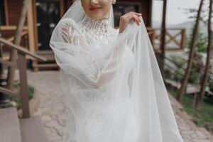 Portrait. The bride in an elegant long dress and veil, holding a bouquet, posing near a wooden railing. Wedding in nature photo