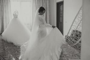 A beautiful brunette bride is getting ready for the wedding in a beautiful boudoir style outfit standing next to her wedding dress by the window. Black and white photo. photo