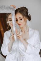 portrait of a luxurious bride in boudoir style near a mirror with reflection. Preparing the bride for the wedding in the morning in a white robe. photo