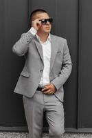 Handsome successful young man wearing gray suit, white shirt, sunglasses and posing against gray wall in modern city. photo