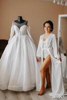 A bride in a lace bodysuit and silk robe with a veil poses in her room next to her wedding dress. Morning of the bride. Preparation. Wedding photography photo