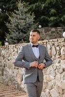The groom in a gray suit adjusts his jacket, poses against the background of a stone wall. Wedding portrait. photo