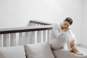 a beautiful girl in white pajamas is sitting on a gray sofa in a wonderful room. Free space photo