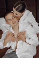 joyful young bride in white pajamas hugging the shoulders of handsome groom in white unbuttoned shirt sitting together in modern hotel room before wedding ceremony photo